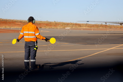 Aircraft worker wearing long sleeve hi-vis tape safety visible shirt earmuffs holding round yellow airplane signal plates both left right hand down giving direction to pilot airplane to stopped     photo