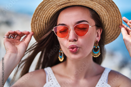 Stylish sensual bright brunette woman with closed eyes wearing a straw hat, big earrings and red glasses enjoying summertime and blowing air kiss