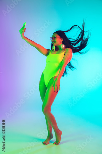 Beautiful young girl taking selfie using smartphone on gradient background in neon. Fit woman in fashionable bodysuit. Facial expression, summer, weekend, beauty, resort concept. Vacations, youth.