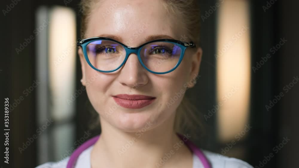 Young Caucasian smiling blonde female student in white medical coat and glasses looking at the camera, closes her eyes and dreams of a bright future. Camera slowly moves back and the frame blurs. Young Caucasian smiling blonde female student in white medical coat and glasses looking at the camera, closes her eyes and dreams of a bright future. Camera slowly moves back and the frame blurs.