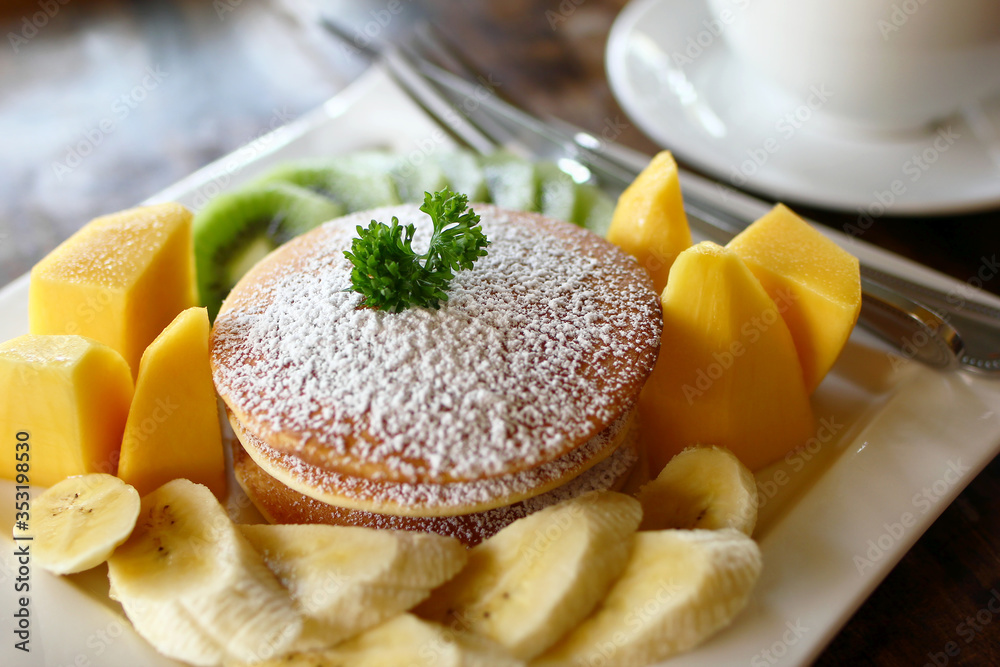 Plate with pancakes with fresh fruits - mango, banana and kiwi - decorated powdered sugar and leaf of mint on a table in cafe with cup of coffee on a background.