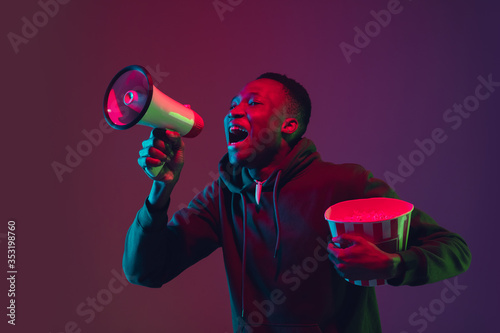 Shouting. African-american man's portrait on gradient studio background in neon light. Beautiful male model with popcorn. Concept of human emotions, facial expression, sales, ad, cinema, entertainment