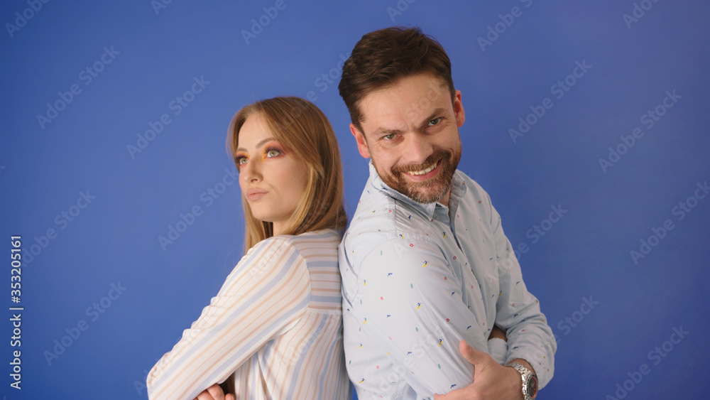 Husband and wife or coworkers. Man with a big smile and angry annoyed woman staying back to back on the blue background.. Husband and wife or coworkers. Man with a big smile and angry annoyed woman staying back to back on the blue background..