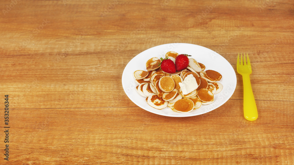 Tiny pancakes with strawberries and butter are on a white plate on a wooden background, next to a yellow plastic fork. Breakfast for children, trendy Breakfast, trendy food. Space for text Tiny pancakes with strawberries and butter are on a white plate on a wooden background, next to a yellow plastic fork. Breakfast for children, trendy Breakfast, trendy food. Space for text