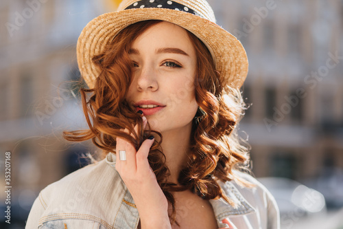 Portrait of interested ginger woman posing on blur background in elegant hat. Outdoor shot of gorgeous female model with red curly hair spending weekend in city.