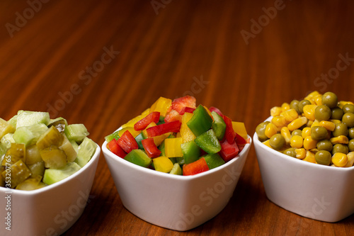 Papryki, ogórki, kukurydza i groszek pokrojone w kostkę do sałatki. Warzywa w białych miskach na brązowym stole gotowe do jedzenia. Świerze kolorowe warzywa na stole w białych miseczkach. 