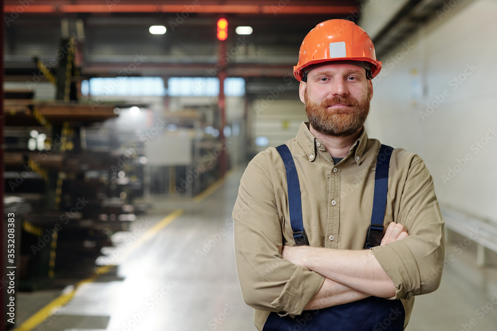 Portrait of content brutal bearded factory man in orange hardhat standing with crossed arms in workshop Portrait of content brutal bearded factory man in orange hardhat standing with crossed arms in workshop