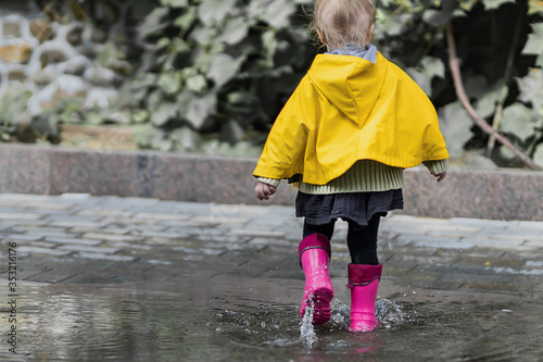 A nice little girl in a yellow raincoat and pink rubber boots jumps on puddles with splashes and rejoices. Park, nature, outdoors. Summer time. Universal Children's Day.