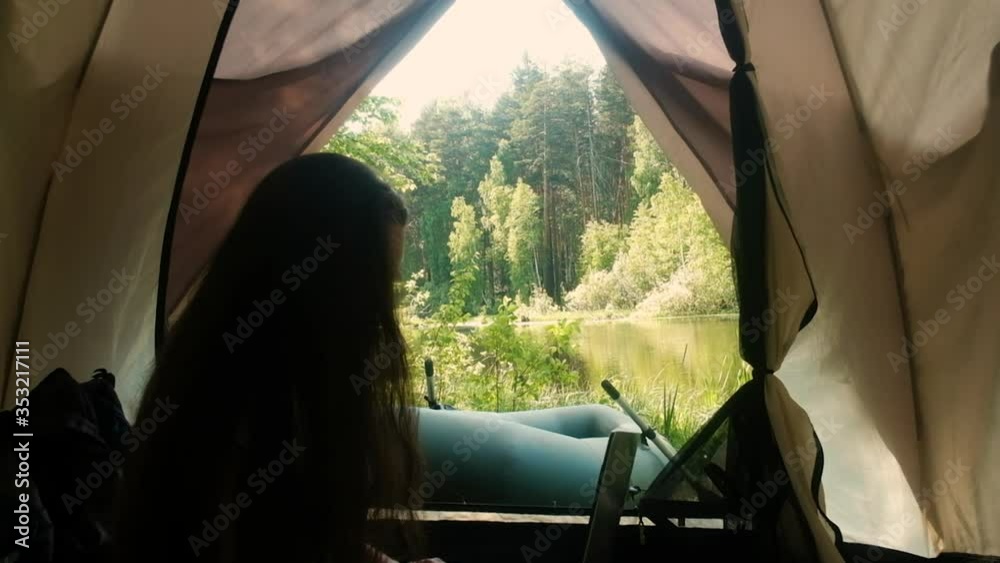 Young woman tourist is sitting in a tent and working on a computer. Camping in the woods. Active lifestyle. Traveling in the morning. Camp by the river. A boat. Internet connection