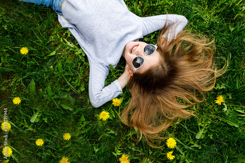 A little girl is smiling lying on the bright green grass with dandelions. Portrait of a happy child on the grass in summer. The view from the top. Space for text