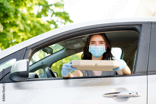 Girl with pizza box in car with medical mask and gloves. Delivery food service drive by car. Safe food delivered according social distance. Tasty pizza with salami, cheese, tomatoes in cardoard box © Rabizo Anatolii