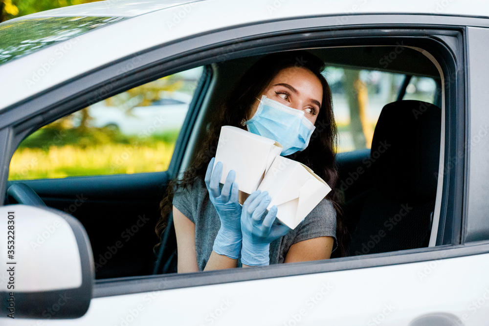 Young attractive girl in car in medical gloves and mask holds wok in box udon noodles with tempuru, shrimps, in hands and smiles. Udon noodles in white box delivery. 2 Wok box udon advertise 1+1. Young attractive girl in car in medical gloves and mask holds wok in box udon noodles with tempuru, shrimps, in hands and smiles. Udon noodles in white box delivery. 2 Wok box udon advertise 1+1.