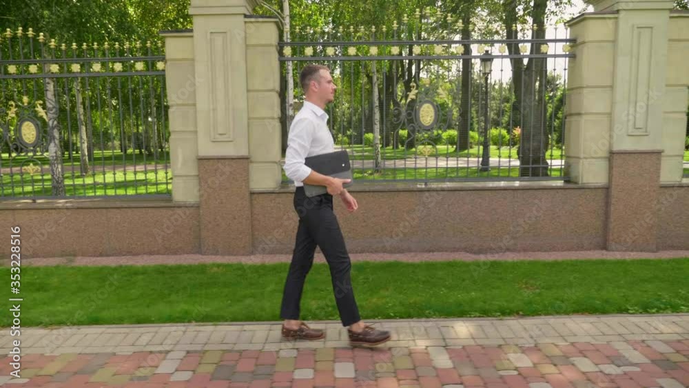 Businessman with laptop in his hand walks through the park. Man freelancer in a white shirt and trousers goes to a meeting with client. 4K footage Businessman with laptop in his hand walks through the park. Man freelancer in a white shirt and trousers goes to a meeting with client. 4K footage