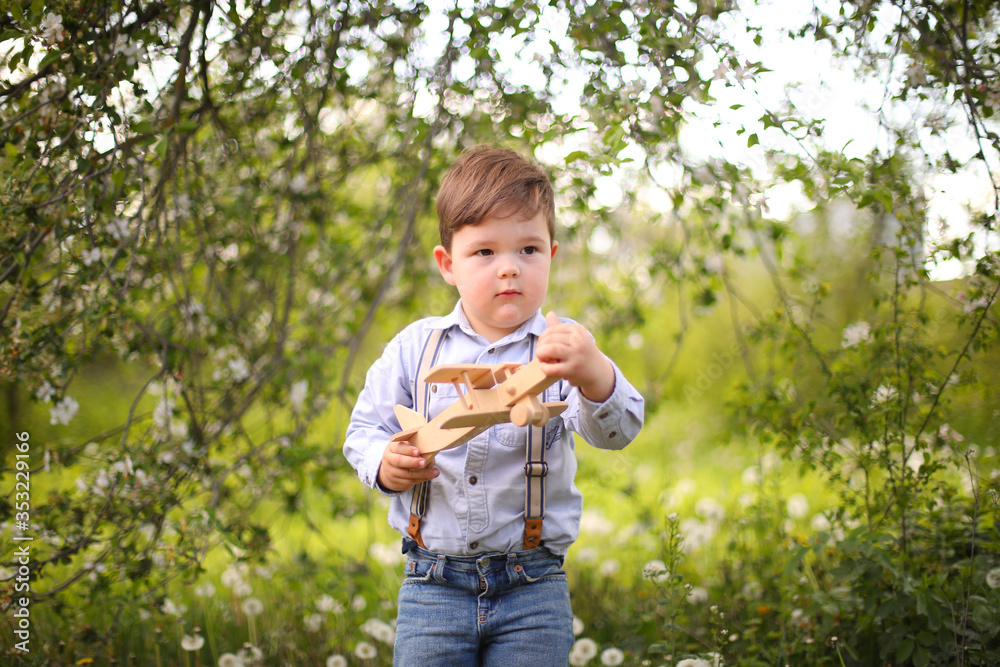 Little cute blond boy playing with a wooden plane in the summer park on the grass on a sunny day, focus on the child, blurred background Little cute blond boy playing with a wooden plane in the summer park on the grass on a sunny day, focus on the child, blurred background