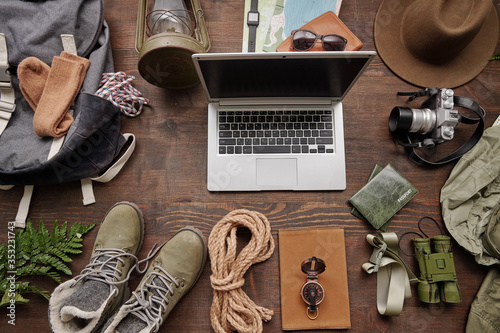 Above view of open laptop among hiking stuff such as lantern, boots, satchel, rope and binoculars on wooden table, flat lay