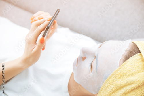 Woman relaxing with a facial textile sheet mask and using a smartphone.