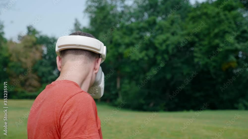 Man in a virtual reality helmet turns his head left and down. Guy in a park in a red T-shirt on background of green trees looks VR glasses. 4K footage Man in a virtual reality helmet turns his head left and down. Guy in a park in a red T-shirt on background of green trees looks VR glasses. 4K footage