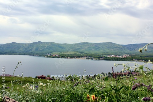 Great panoramic view. A lagoon with a clean mountain lake in the midst of majestic mountains in a haze of fog. Green grass, yellow and blue flowers in the foreground