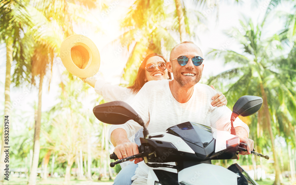 Laughing happy couple travelers riding motorbike during their tropical vacation in Thailand under palm trees. Woman raised hands up with hat. - obrazy, fototapety, plakaty 