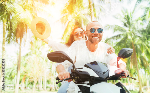 Laughing happy couple travelers riding motorbike during their tropical vacation in Thailand under palm trees. Woman raised hands up with hat.
