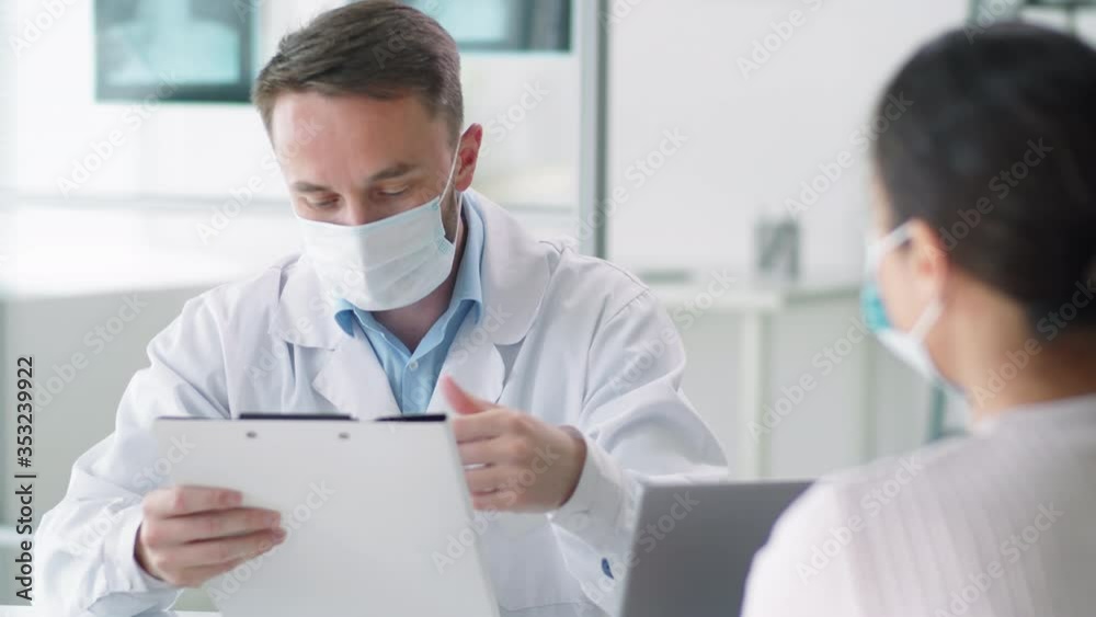 Male physician in protective face mask and lab coat checking medical history of female patient and talking with her during consultation in clinic. Coronavirus outbreak concept Male physician in protective face mask and lab coat checking medical history of female patient and talking with her during consultation in clinic. Coronavirus outbreak concept