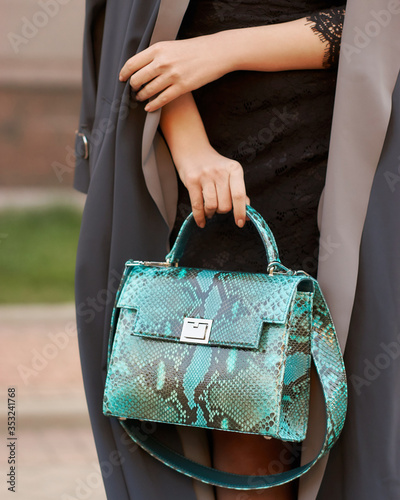 Woman holding green reptile leather handbag close-up. Fashion details. Beige colors. Outdoors. Black stone wall background