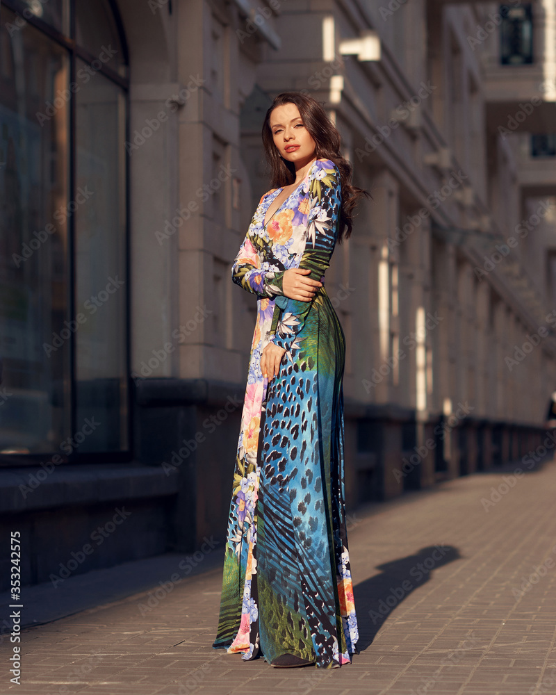 Elegant woman wearing long colorful evening dress and walking city street at sunset light. Pretty female model with long wavy hair standing and posing