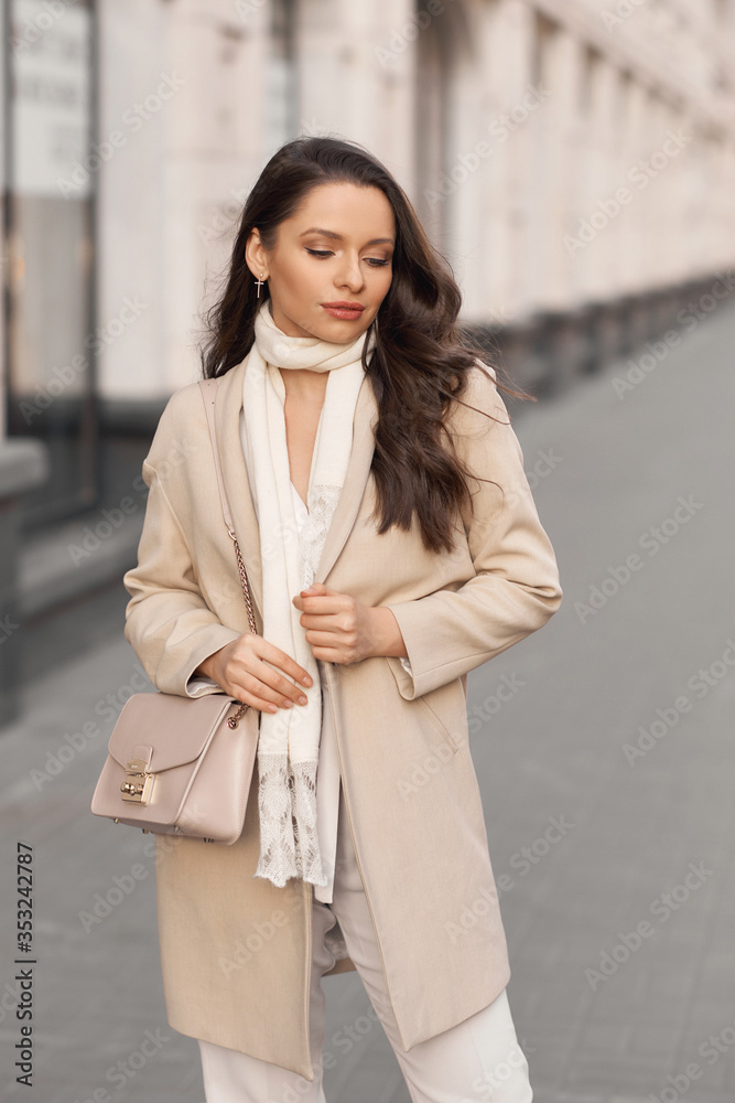 Outdor portrait. Elegant woman in beige jacket with white scarf walking city street holding leather handbag. Pretty female model with long wavy brunette hair and perfect makeup