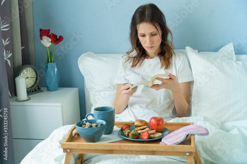 Young beautiful woman reading book at morning in bed with breakfast on tray.