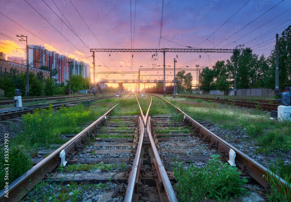 Obraz premium Railway station with freight trains at colorful sunset. Railroad in summer. Heavy industry. Industrial landscape with train, green grass, railway platform, purple sky with pink clouds. Transportation