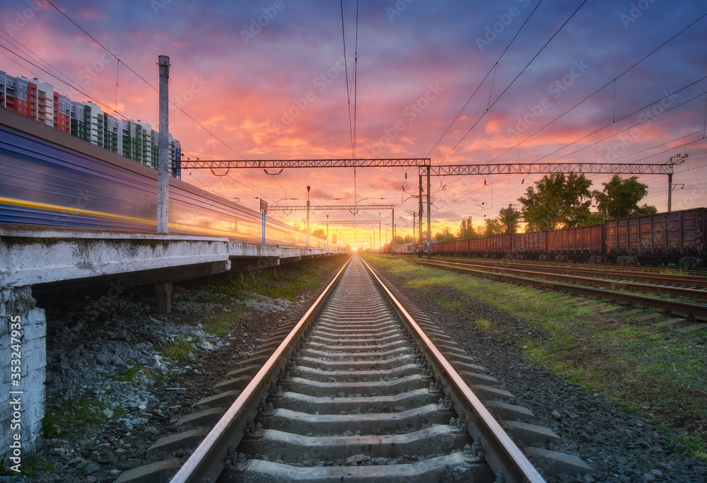 Railway station with blurred high speed train and freight train at colorful sunset. Railroad in summer. Industrial landscape with moving train, railway platform, sky with pink clouds. Transportation Railway station with blurred high speed train and freight train at colorful sunset. Railroad in summer. Industrial landscape with moving train, railway platform, sky with pink clouds. Transportation