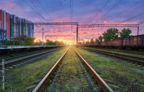 Railway station with freight trains at colorful sunset. Railroad in summer. Heavy industry. Industrial landscape with train, green grass, railway platform, purple sky with pink clouds. Transportation