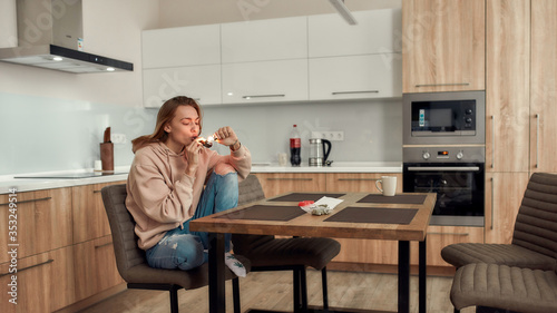 Stress Relief. Young caucasian woman lighting cannabis in the bowl of metal pipe while sitting in the kitchen. Buds in a plastic bag and red marijuana grinder on the table