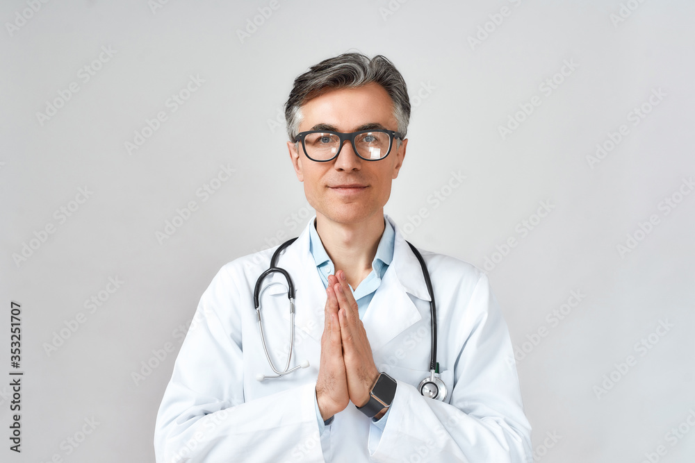 Senior professional male doctor or physician in white coat with stethoscope around neck praying, looking at camera while standing against grey background Senior professional male doctor or physician in white coat with stethoscope around neck praying, looking at camera while standing against grey background