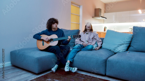 Young girl smoking marijuana from glass water pipe, exhaling the smoke, resting with boyfriend on the sofa at home. Guy playing guitar