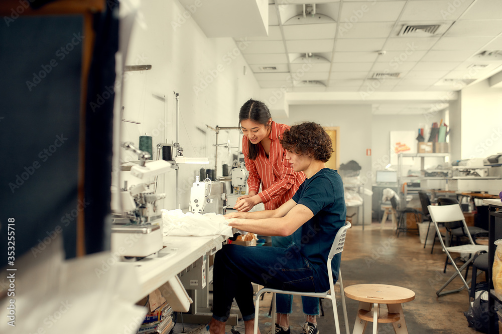 Obraz premium The Joy of Crafting Best. Young man, clothing designer sitting and sewing. His female colleague helping him. Dressmaker working on the sewing machine. Tailor making a garment in his workplace