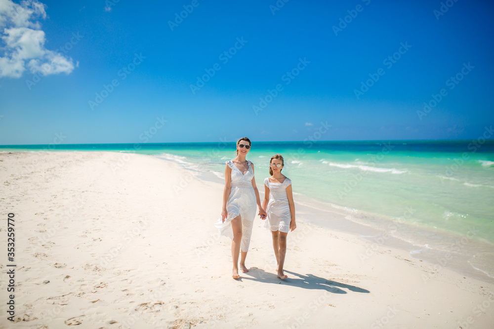 Mother and daughter in white dresses walking on the beach. Family by the ocean in sunglasses.