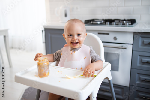 baby girl sits in the kitchen in a feeding chair with a spoon in her hands and smiles photo