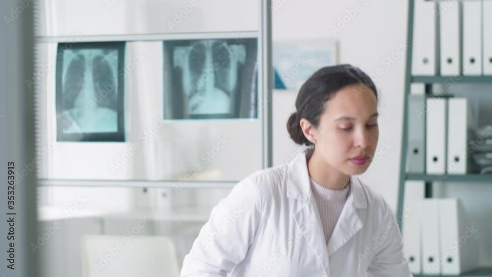 Beautiful mixed-raced female doctor in lab coat holding clipboard, going into medical office, sitting at desk and working on laptop Beautiful mixed-raced female doctor in lab coat holding clipboard, going into medical office, sitting at desk and working on laptop