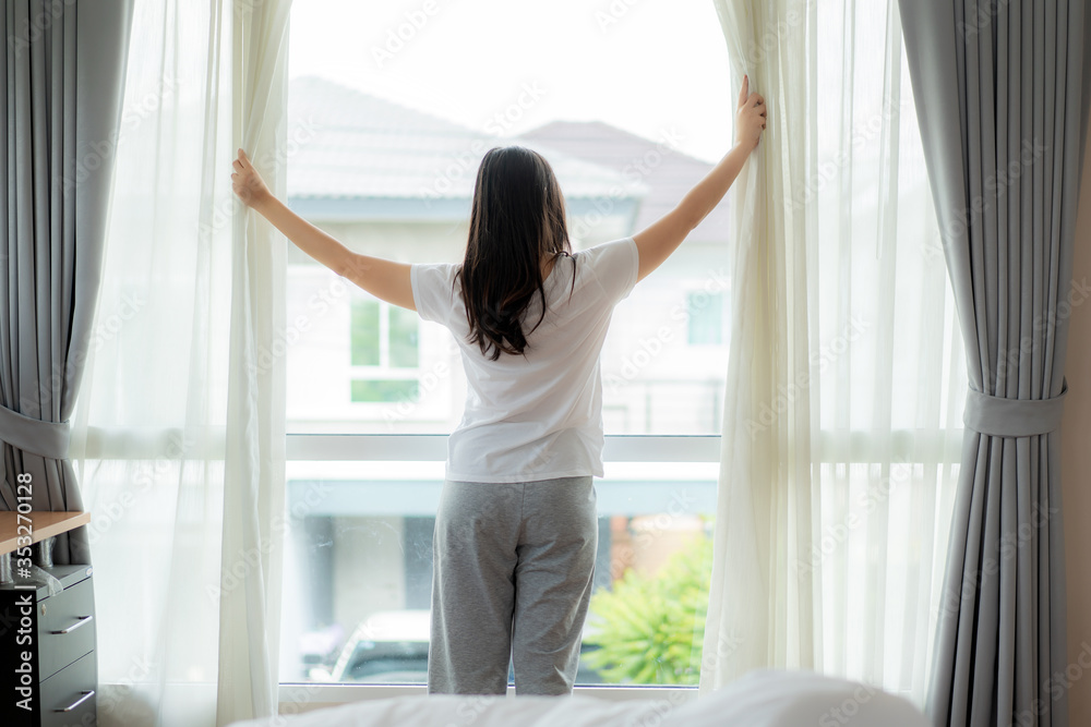 Rear view of Asian woman waking up in her bed fully rested opening window curtains and looking through the window in bedroom at home. Rear view of Asian woman waking up in her bed fully rested opening window curtains and looking through the window in bedroom at home.