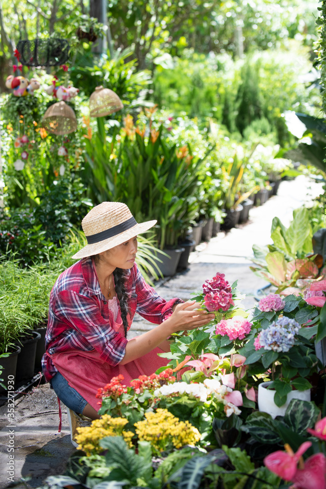 Young woman at a nursery holding flower in her hands as she kneels in the walkway between plants with a basket of fresh white flowers for sale