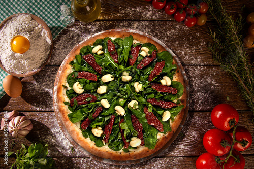 Tasty arugula and dried tomato pizza and cooking ingredients tomatoes basil on wood background. Top view of arugula pizza
