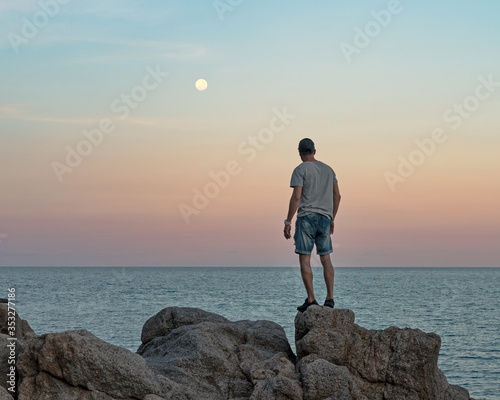 Man in casual clothes stands on the rocks by the sea at sunset and watching the full moon raised 