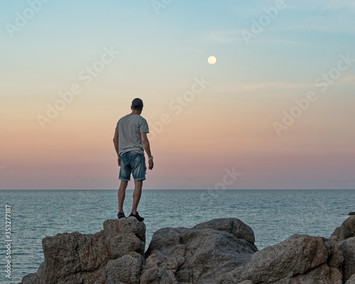 Man in casual clothes stands on the rocks by the sea at sunset and watching the full moon raised 