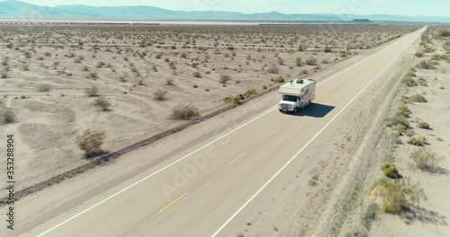 AERIAL - A drone follows a brown camper van from the front in the middle of the Mojave Desert on route 66 on a clear blue day.