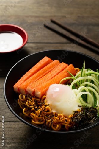 Spicy instant noodle with crab stick and onsen egg and cucumber in black bowl on wooden table.
