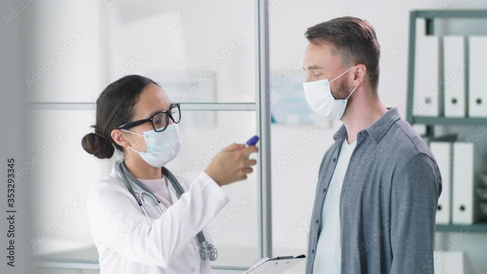 Female doctor in protective face mask scanning forehead of male patient with digital infrared thermometer and writing down measurement on clipboard during coronavirus testing in clinic Female doctor in protective face mask scanning forehead of male patient with digital infrared thermometer and writing down measurement on clipboard during coronavirus testing in clinic