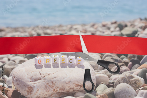 End of quarantine covid-19. The beginning of travel, the opening of borders. Sale of airline tickets. Scissors cut a red ribbon overlooking sea stones pebbles with the inscription Greece