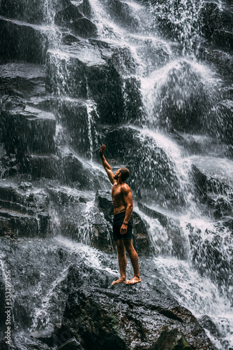 Back view of a man standing by waterfalls with arms outstretched. Man at waterfall raising his hands in feeling closer to nature. Man at the waterfall. Travel to Bali. Man on the background of nature