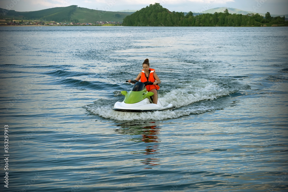A young woman in a life jacket rides a water bike on a lake against the backdrop of the shore in summer.